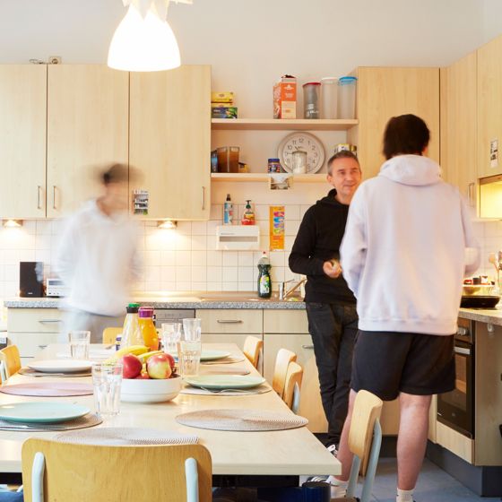 Drei Bewohner in der Küche, im Hintergrund die Küchenzeile, im Vordergrund ein gedeckter Tisch